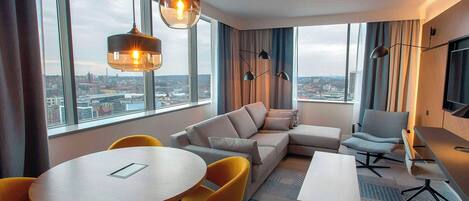 A modern hotel room with a large window, a sofa, a dining table with chairs, and a small desk.
