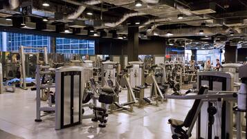 A gym with various exercise machines, large windows, and a high ceiling with exposed ductwork.