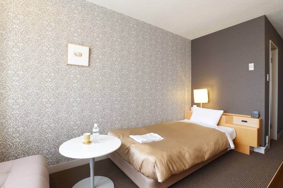 A hotel room with a patterned wallpaper, a bed with a beige bedspread, a bedside table with a lamp, and a small round table with a bottle and a glass.