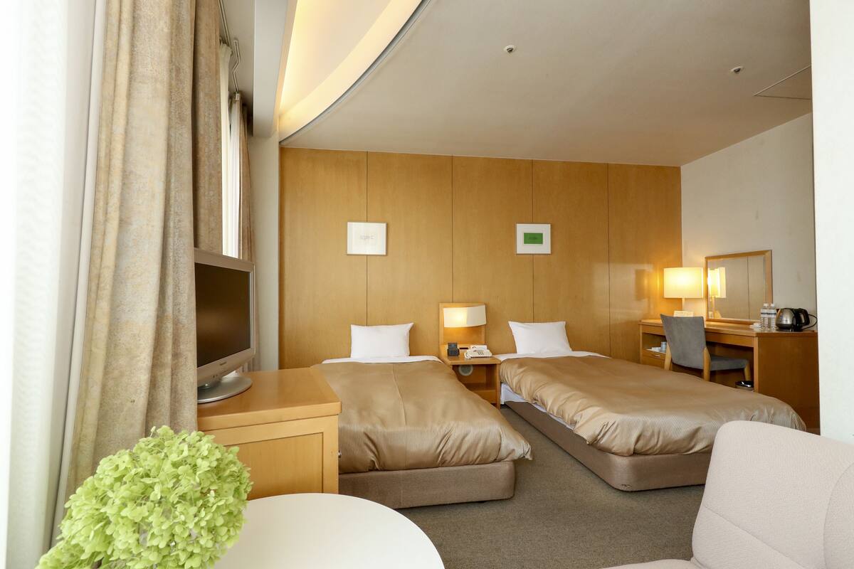 A hotel room with two beds, a desk, a chair, a TV, and a small table with a lamp.