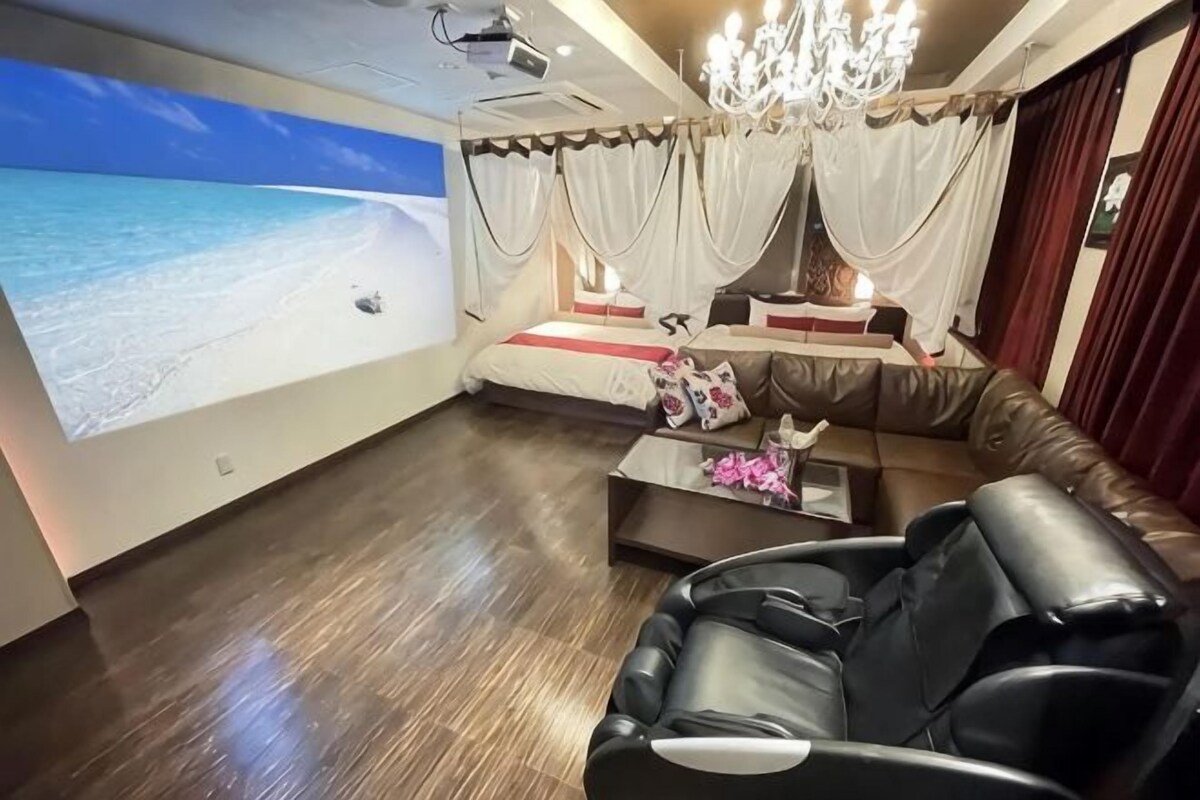 Una habitación con una pantalla grande mostrando una escena de playa, un sofá reclinable, una silla de masajes y una araña.