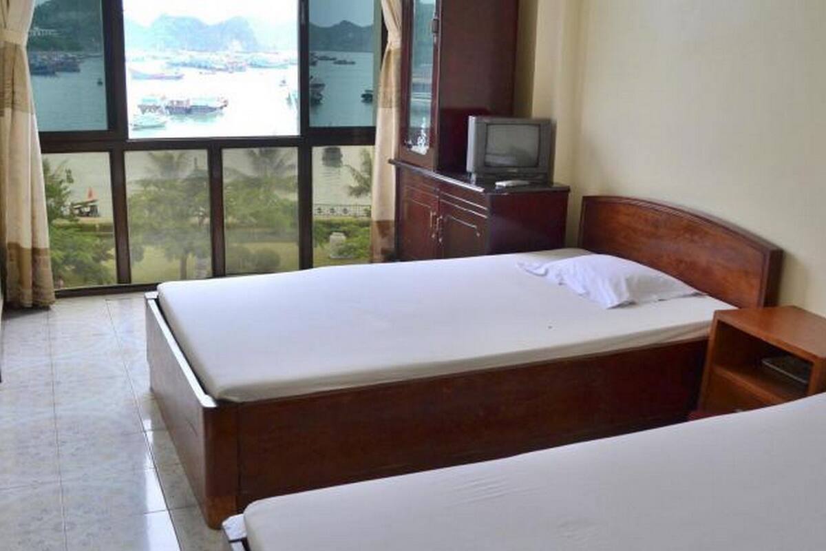 Habitación de hotel con dos camas, cabecero de madera, televisor sobre un mueble de madera y vistas a barcos en el agua a través de amplias ventanas.