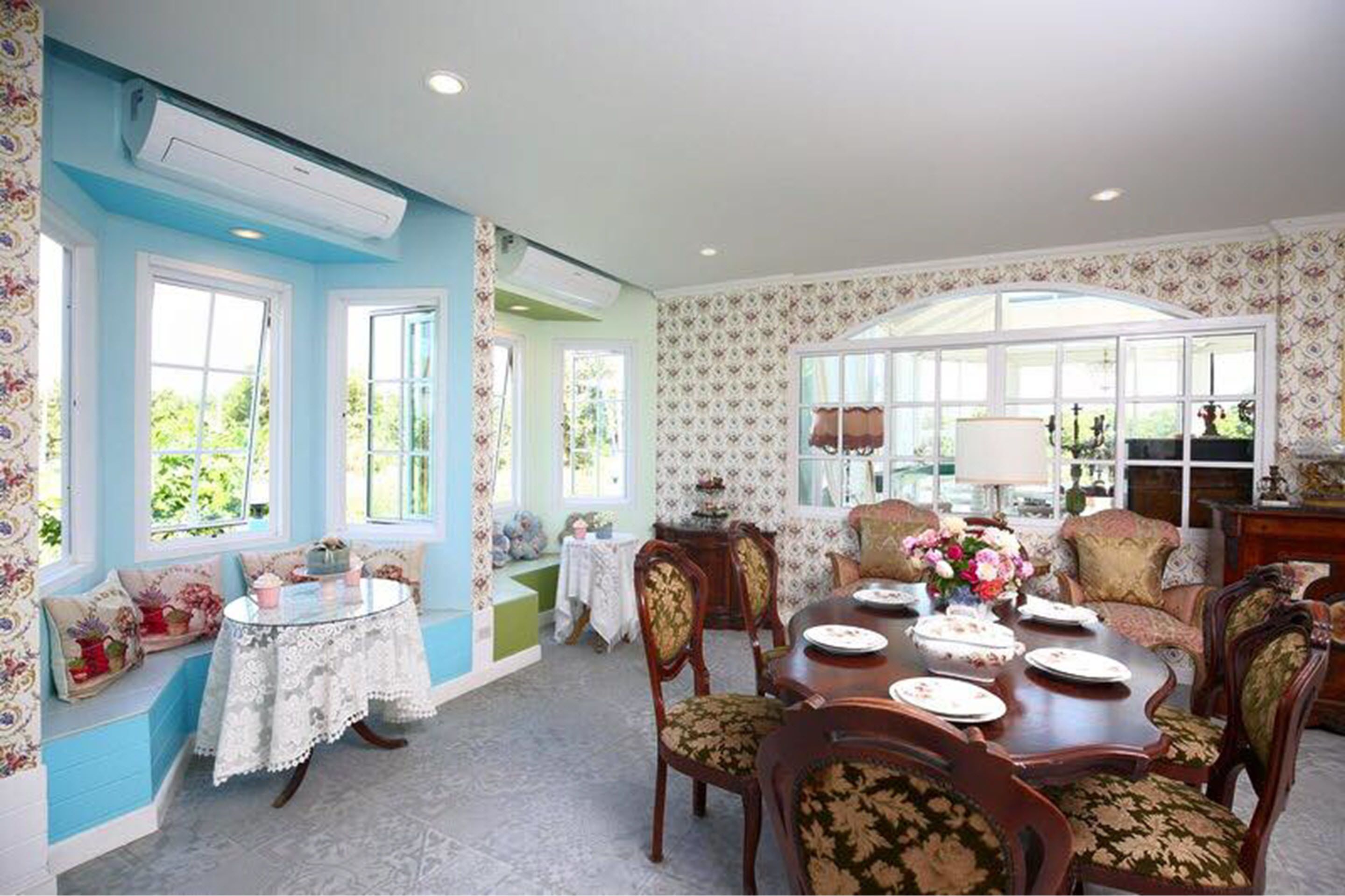A dining area with a round table set for four, surrounded by upholstered chairs with floral upholstery. There are two windows with white frames and floral curtains on the left side, and a large arched window with a white frame on the right.