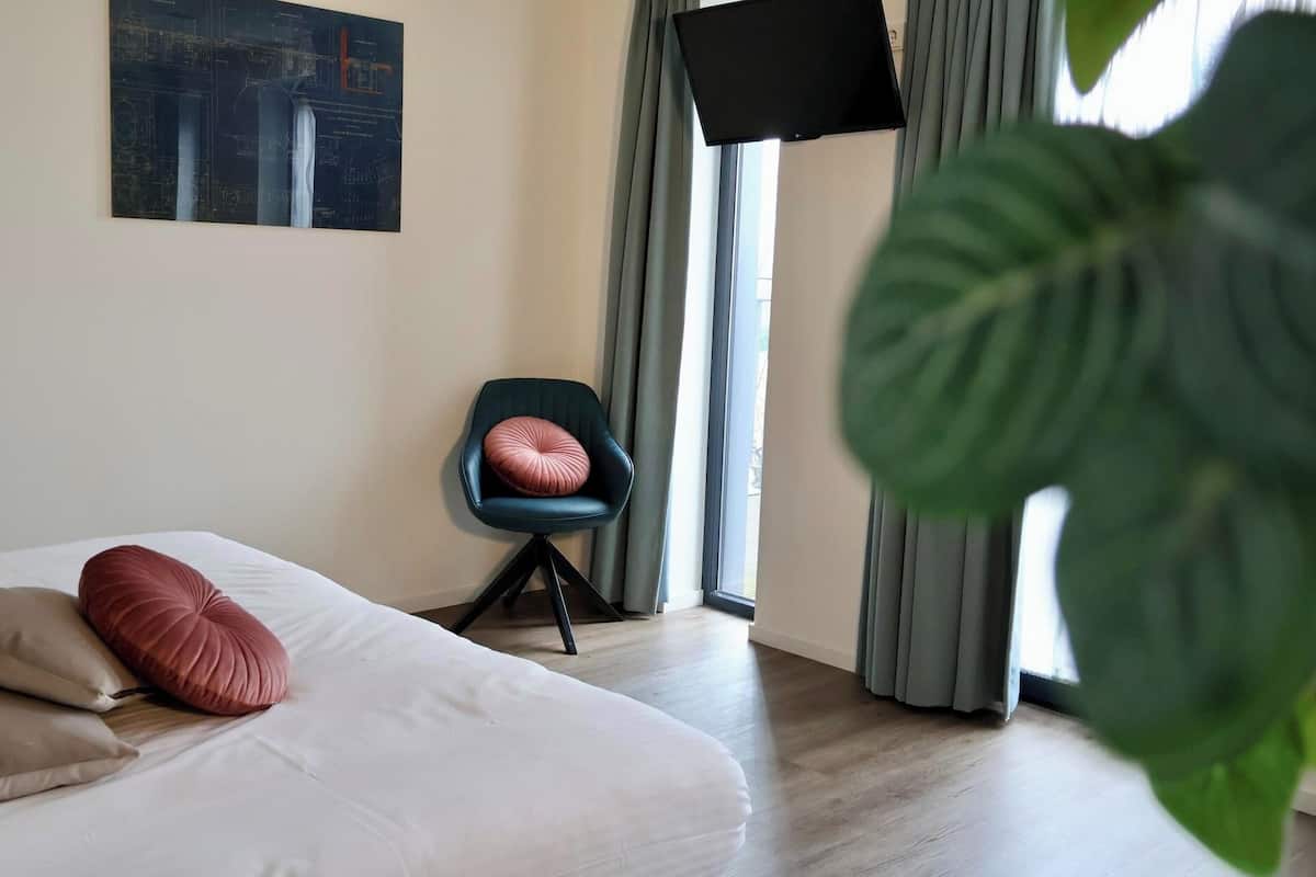 A hotel room with a bed, a chair, a flat-screen TV, and a large window with curtains.