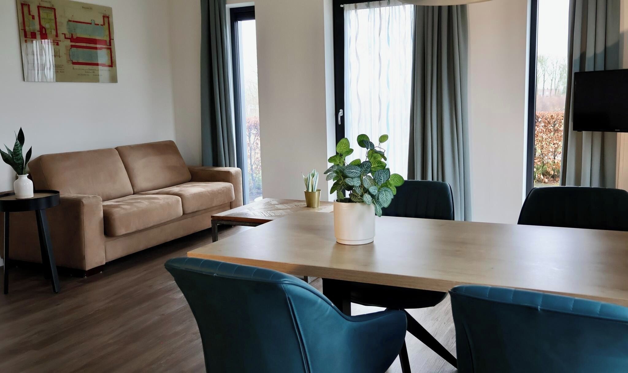 Ein modernes Wohnzimmer mit einem beigefarbenen Sofa, einem Holzesstisch und zwei blauen Stühlen. Zwei Fenster mit Vorhängen, ein kleiner Beistelltisch mit einer Pflanze und ein an der Wand befestigtes Fernsehgerät.