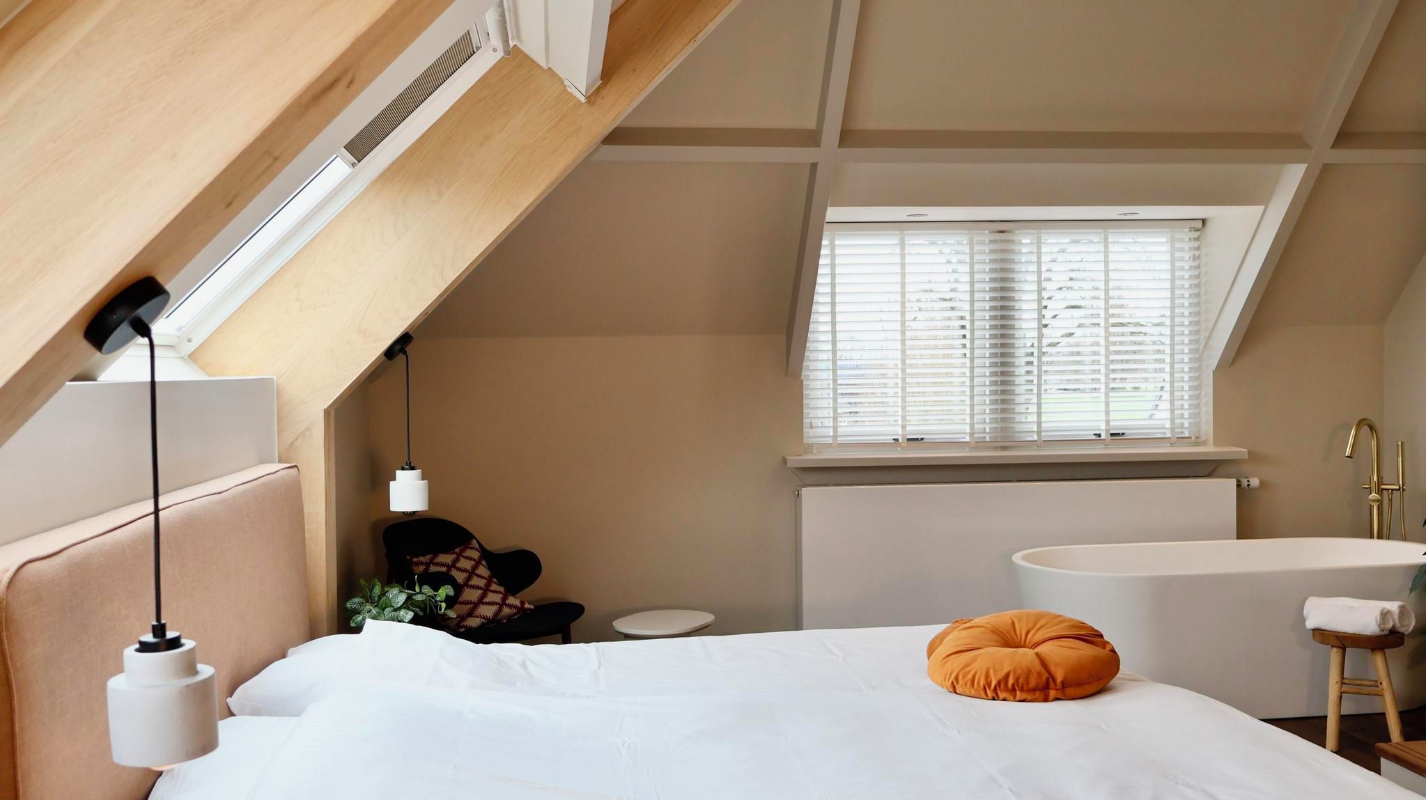 Un dormitorio con techo inclinado, un ventanal con persianas, una cama blanca con almohada blanca, una almohada redonda anaranjada, una mesita de noche y un radiador blanco.