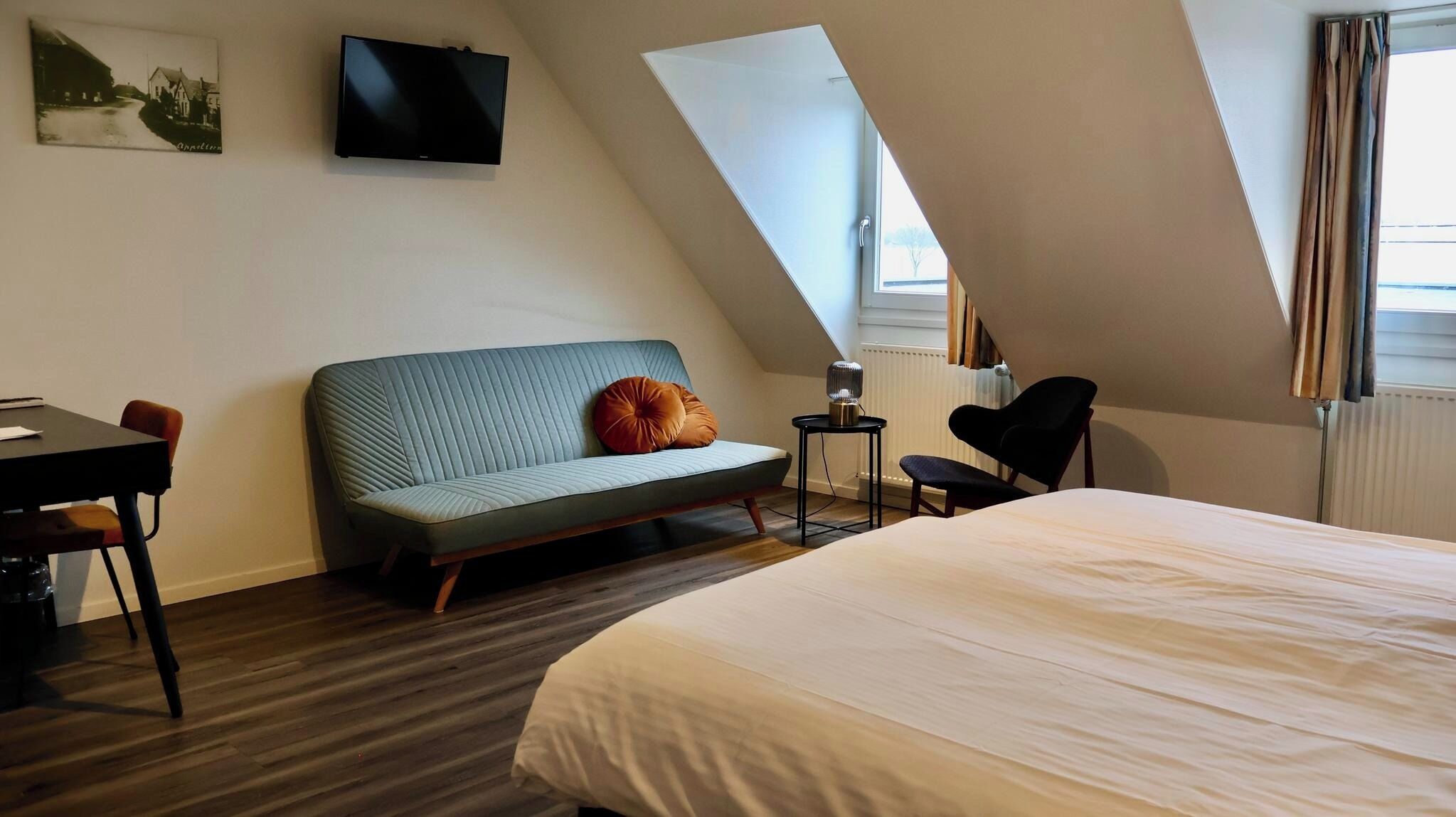 Quarto de hotel moderno com teto estilo loft, uma cama grande, sofá, escrivaninha e televisão fixada na parede.
