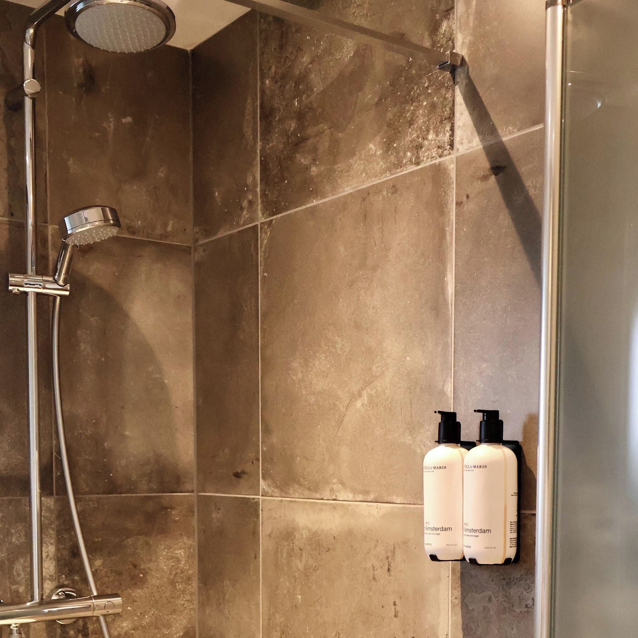 Zona de ducha con ducha de mano y grifo empotrado en la pared. Hay dos botellas de Stella Maris Amsterdam en la pared.