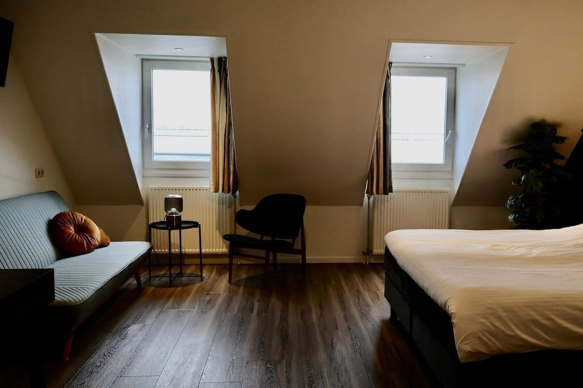 A bedroom with a bed, a chair, a small table, and a sofa. There are two windows with curtains.