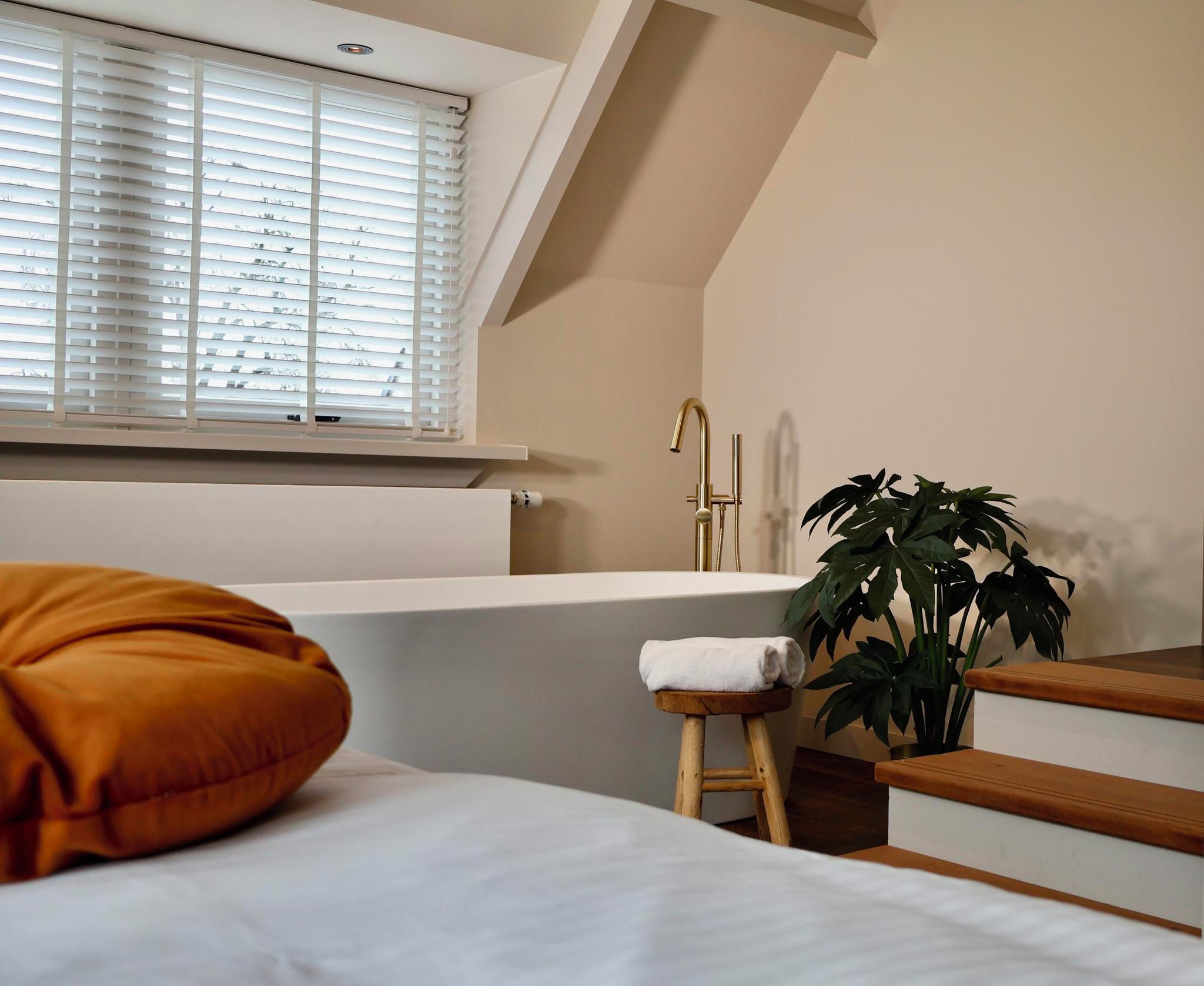 A bedroom with a large window, a bathtub, a plant, and a wooden stool.