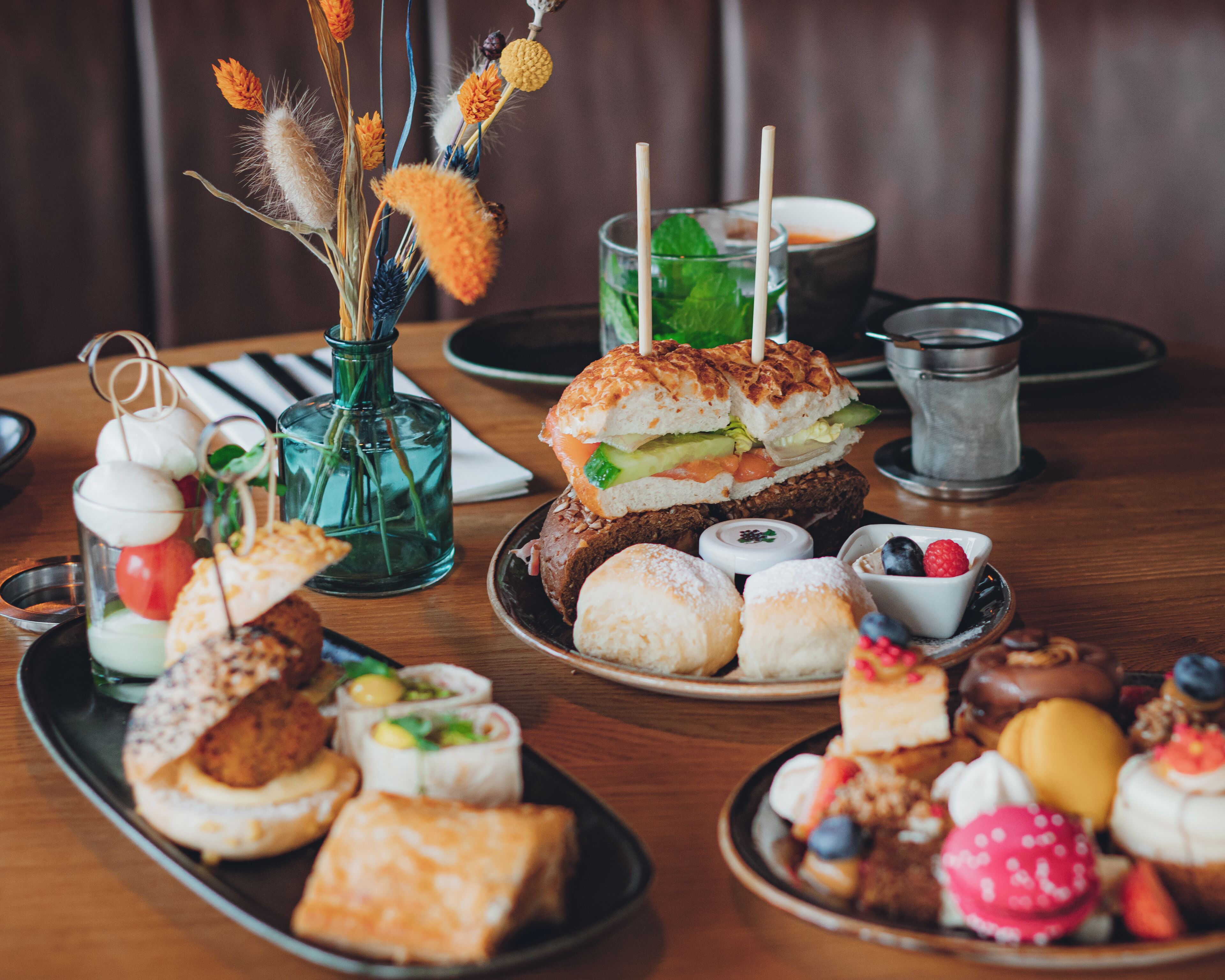 Variedad de pasteles y sándwiches sobre una mesa de madera, con un jarrón de flores secas al fondo.