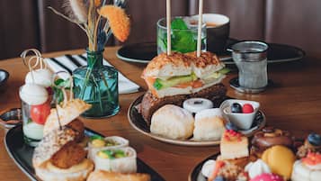 Variedad de pasteles y sándwiches sobre una mesa de madera, con un jarrón de flores secas al fondo.