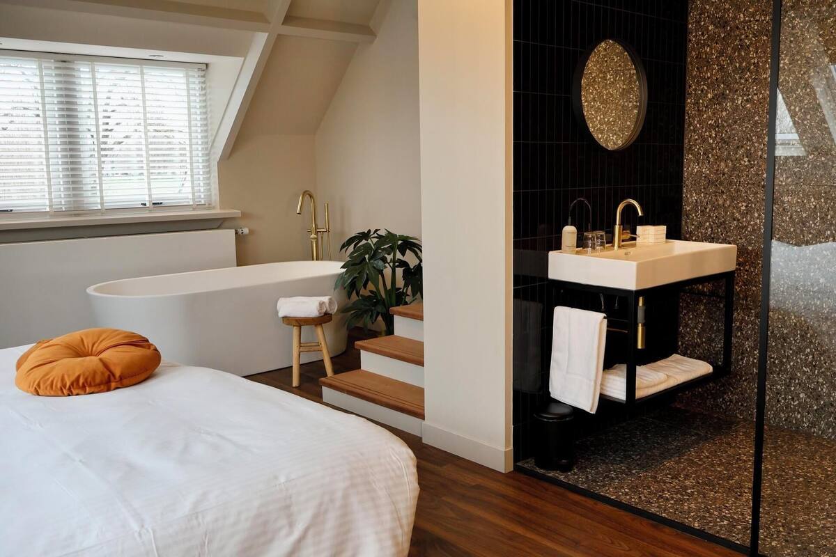 A bedroom with a large bed, a window with blinds, a plant, and a view of a bathroom with a bathtub, sink, and mirror.