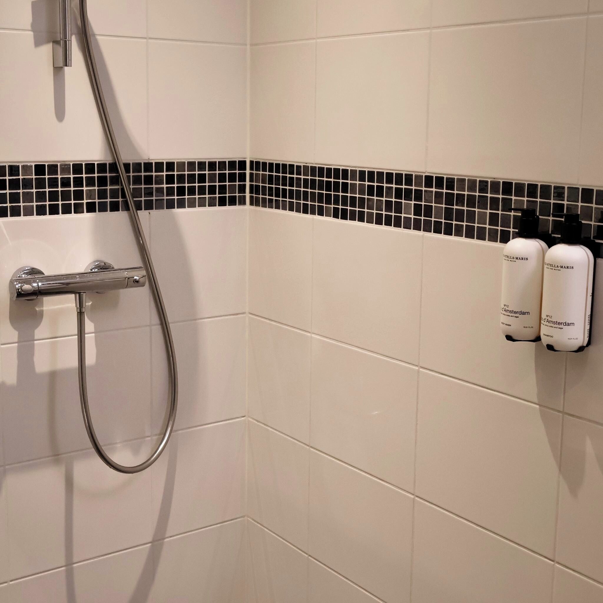Un baño con azulejos blancos, una ducha y dos botellas de champú en la pared.