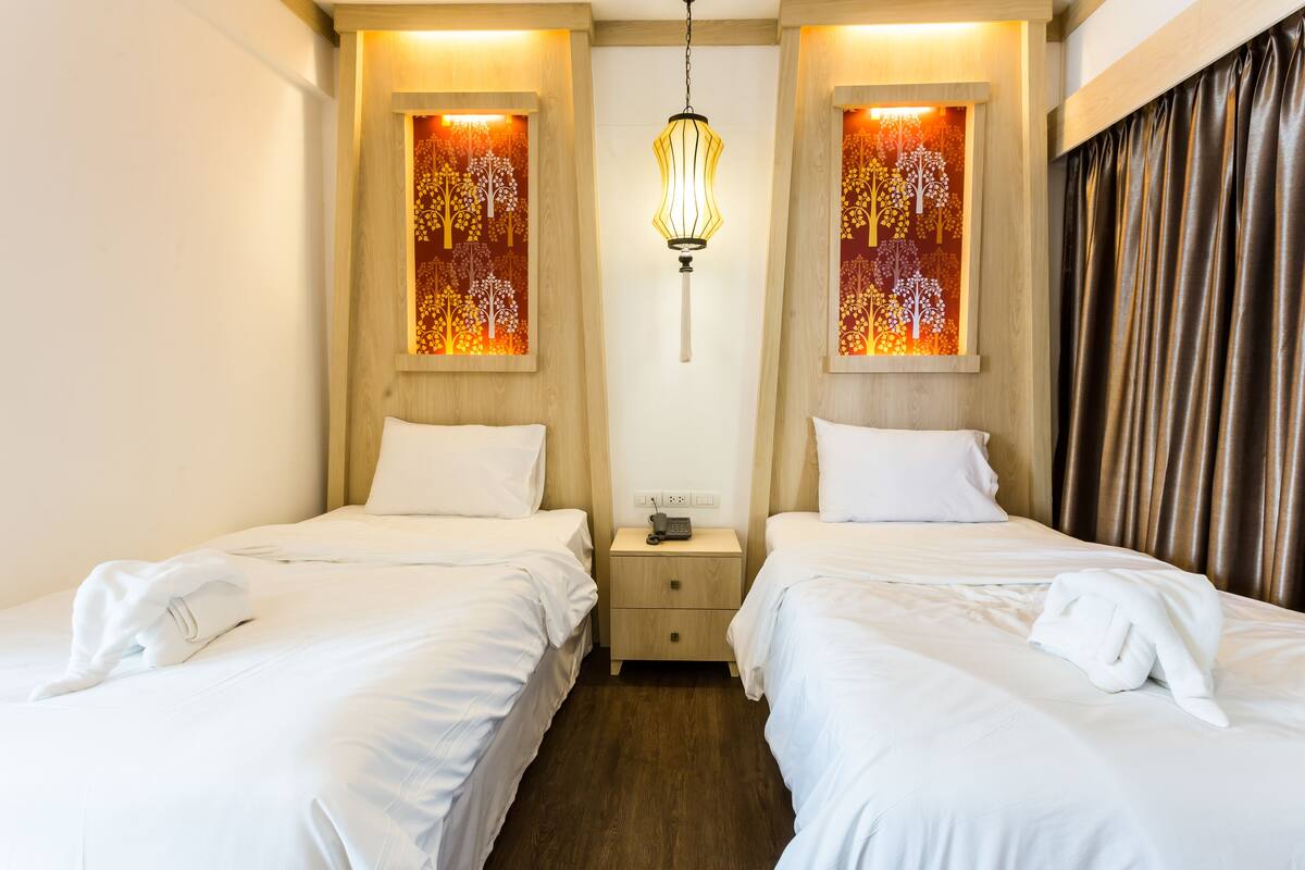 Habitación de hotel con dos camas, una mesita de noche con lámpara y un muro con estampado de árbol.