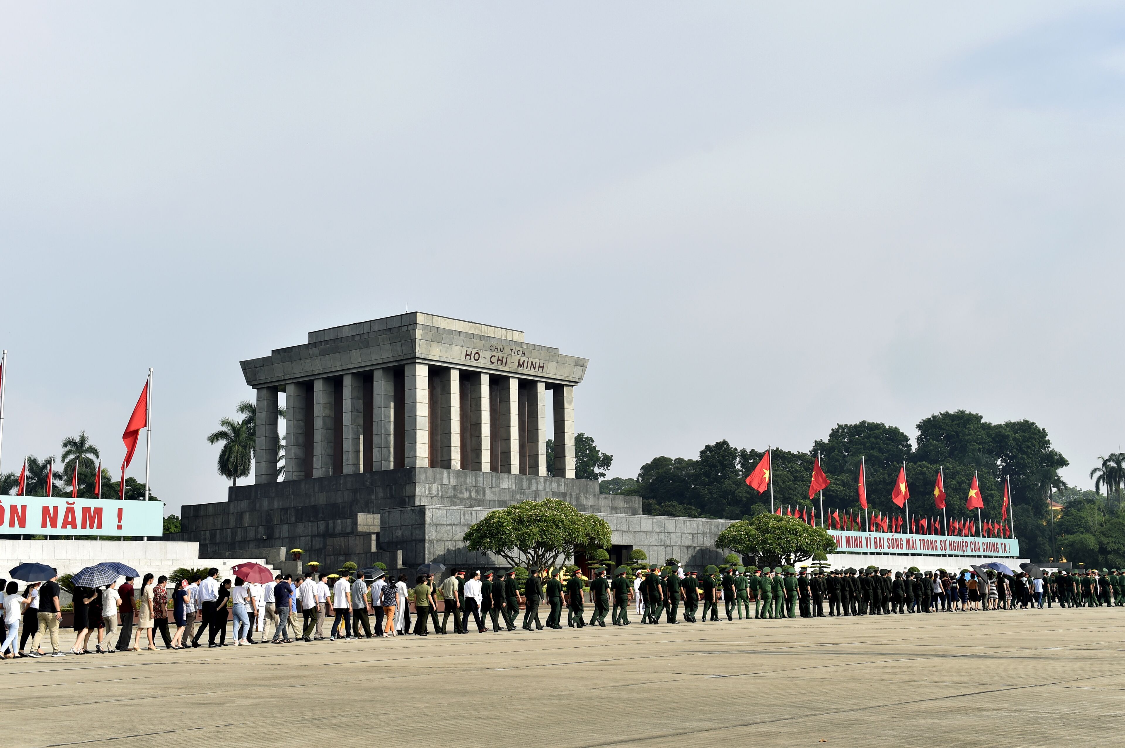 Une vaste place publique, avec un bâtiment imposant en arrière-plan, une foule de personnes et plusieurs drapeaux.