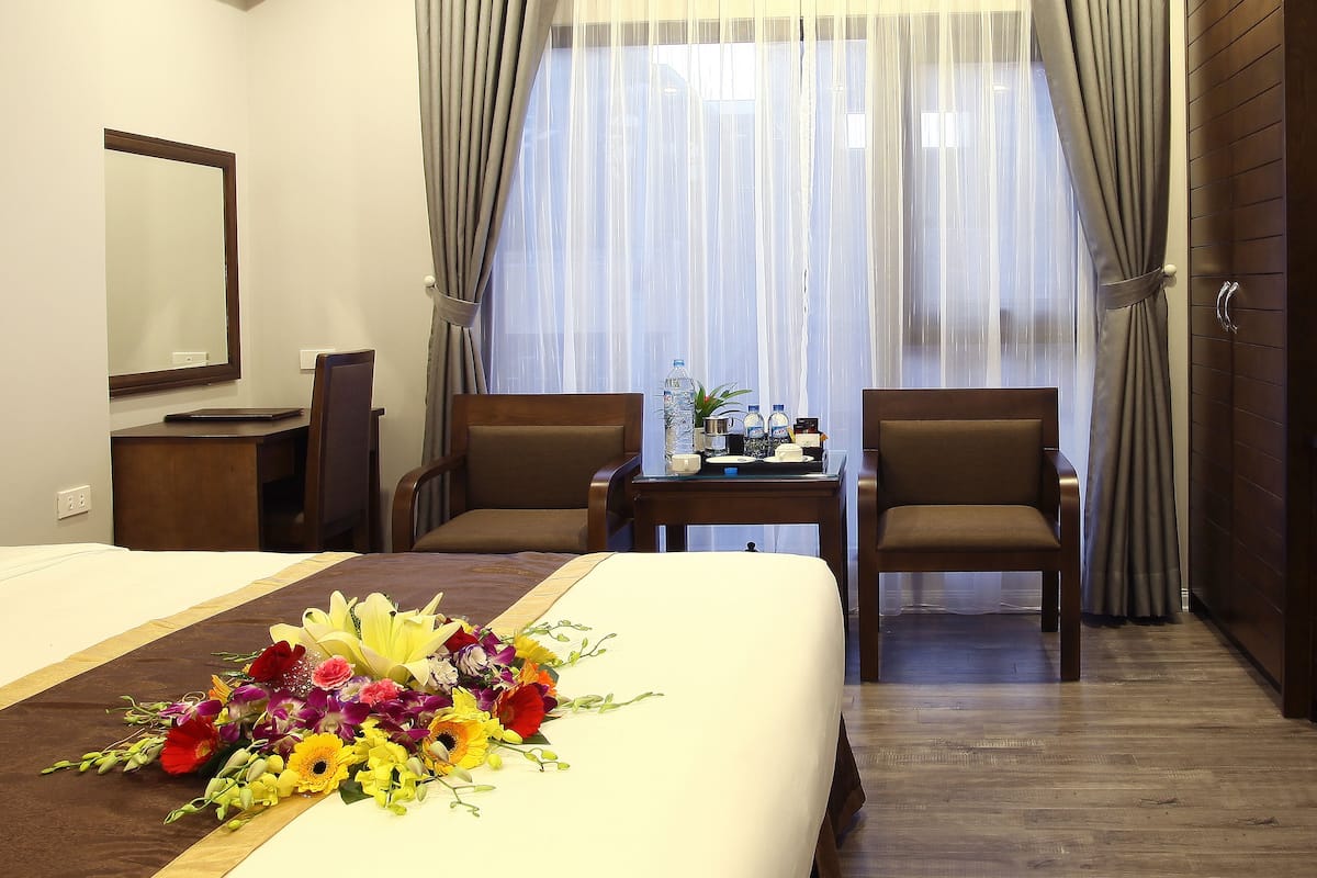 Habitación de hotel con cama, dos sillones, una mesita con una botella de agua, una planta y un espejo en la pared.