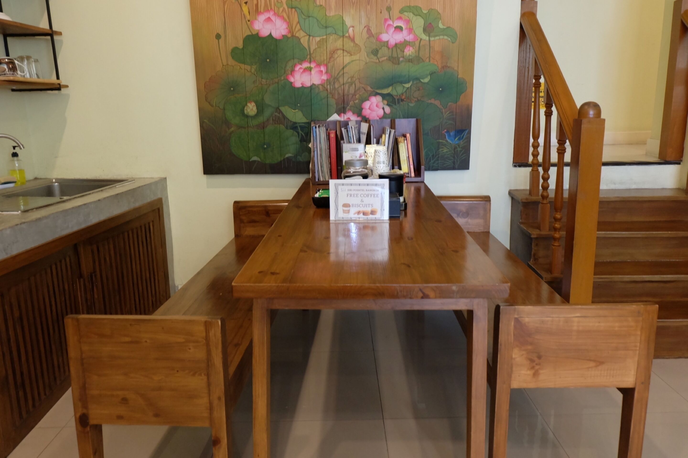 A wooden dining table with chairs, a painting of flowers on the wall, and a sign offering free coffee and biscuits.