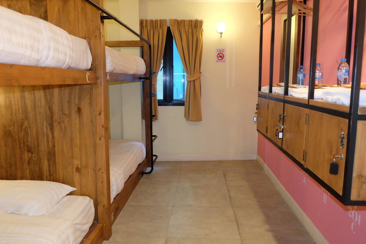 Um dormitório com beliches de madeira, uma janela com cortinas, uma luminária na parede e uma garrafa de água no lado direito.
