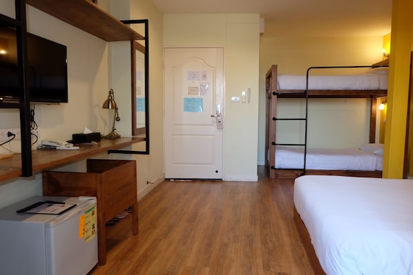 Una habitación de hotel con piso de madera, un escritorio con lámpara, un televisor, camas individuales superpuestas y un refrigerador.