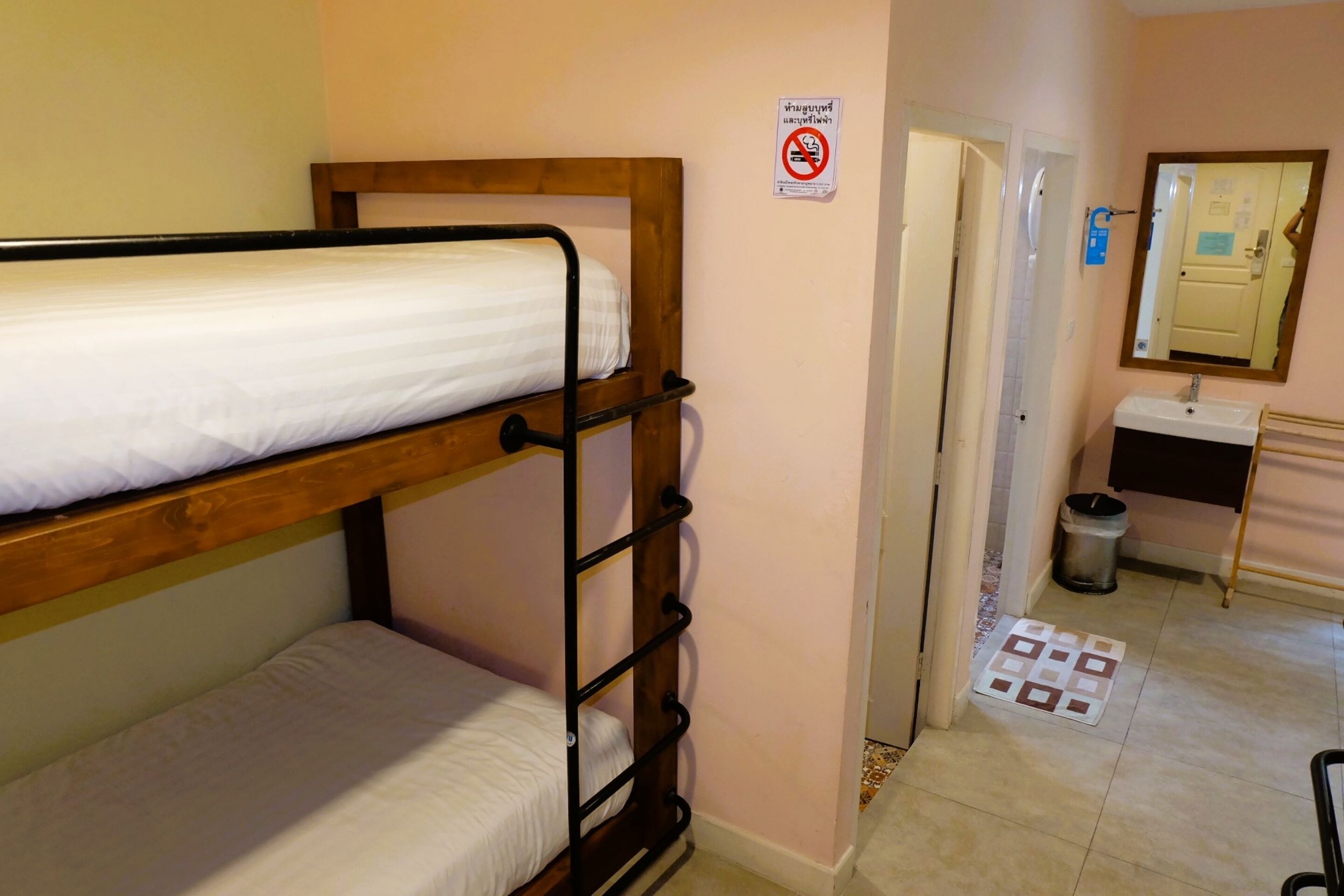A bunk bed with a wooden frame and white bedding, a bathroom with a sink and mirror, and a no-smoking sign on the wall.
