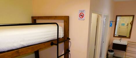 Un lit superposé avec une structure en bois et du linge de lit blanc, une salle de bain avec un lavabo et un miroir, et un panneau «
interdiction de fumer
» sur le mur.