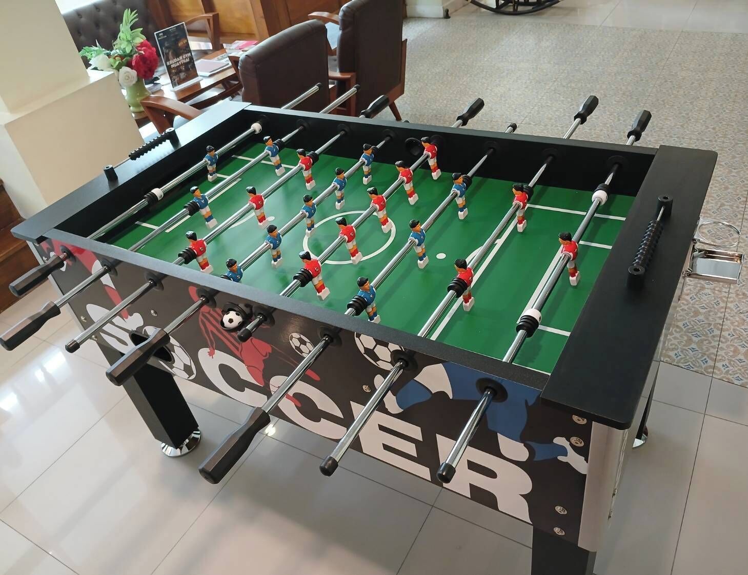 A foosball table with a soccer theme, located in a room with a tiled floor and a sofa in the background.