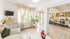 Un salon moderne avec un canapé, une table basse agrémentée d’une plante, un téléviseur fixé au mur et une cuisine visible à travers une porte ouverte.