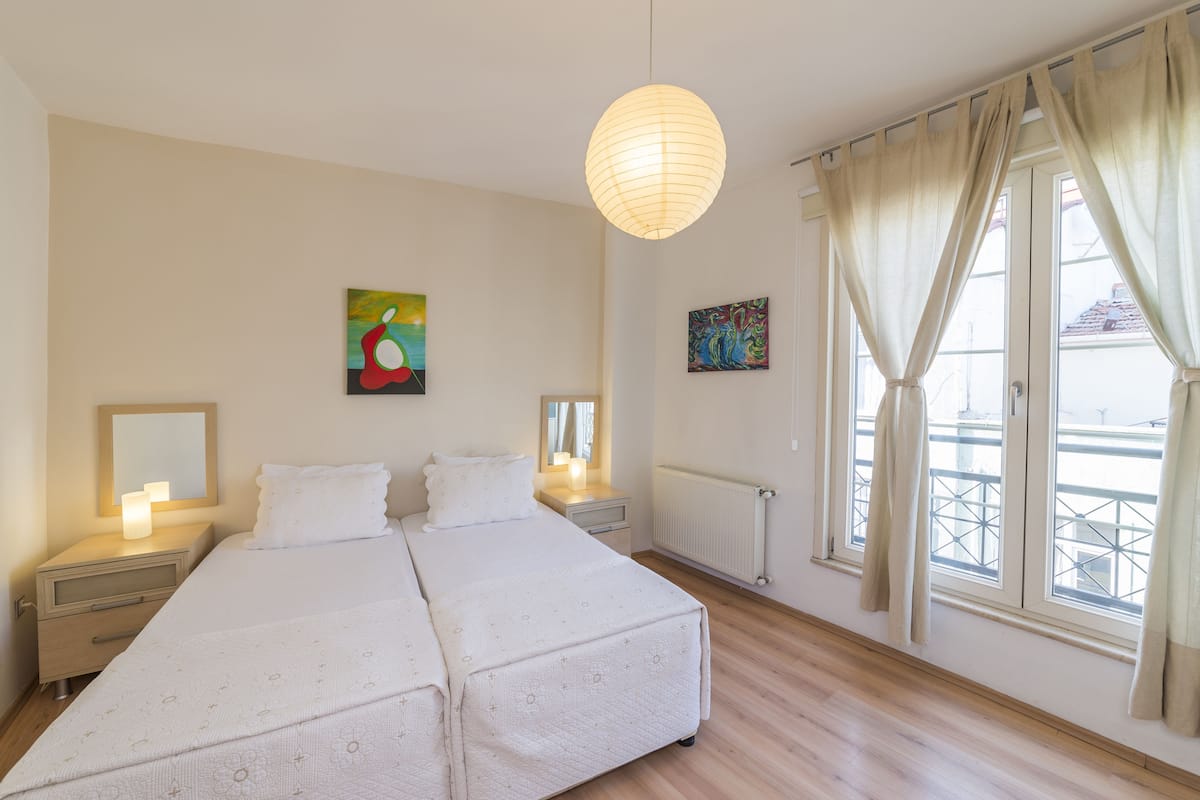 A bedroom with two beds, a nightstand with a lamp, a radiator, and a window with curtains.