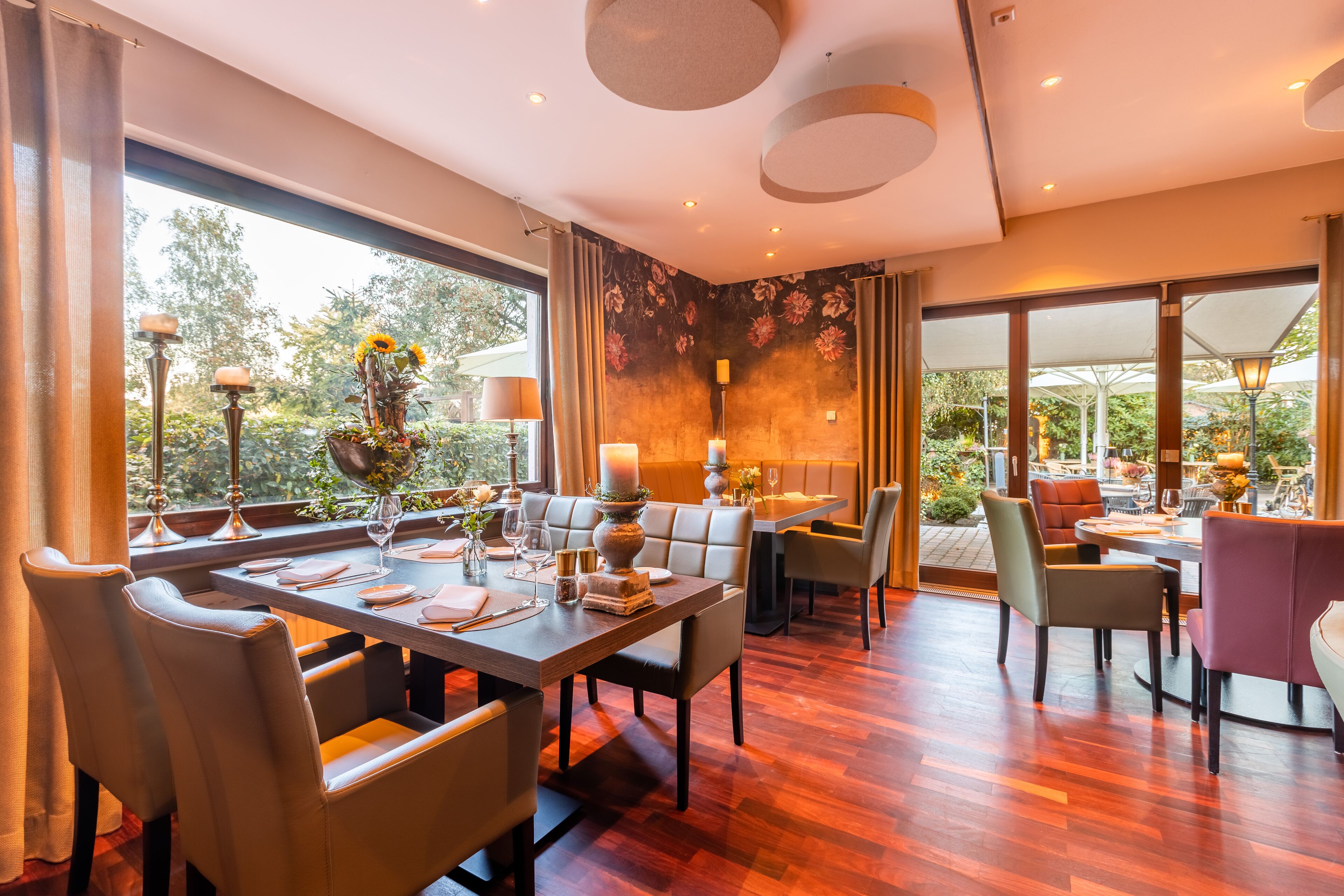 A dining area with a long table set for four, surrounded by chairs, with a view of the outdoors through large windows.