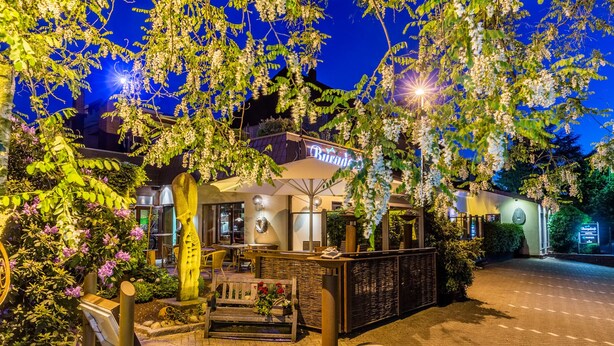 L’entrée d’un hôtel, avec un espace extérieur abrité où sont disposées des tables et des chaises, un grand parasol et un panneau indicateur.