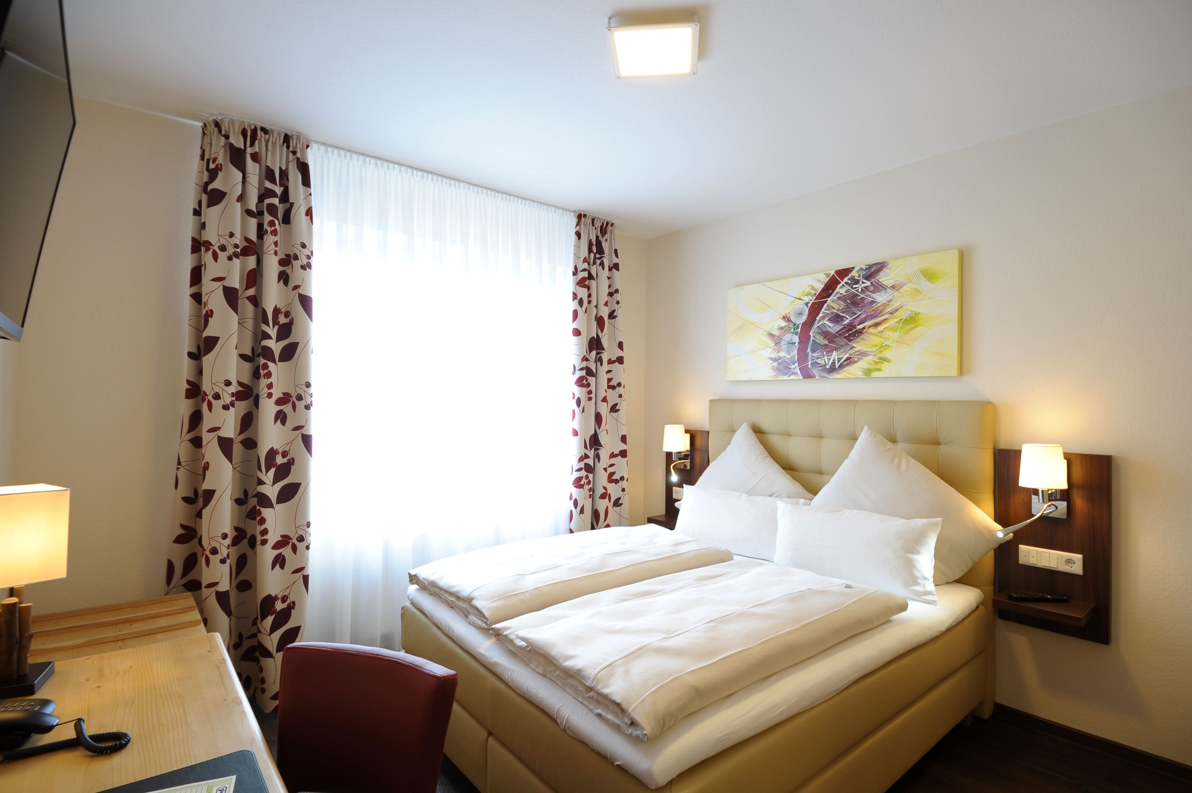 A hotel room with a double bed, a desk with a lamp, a chair, and a painting on the wall.