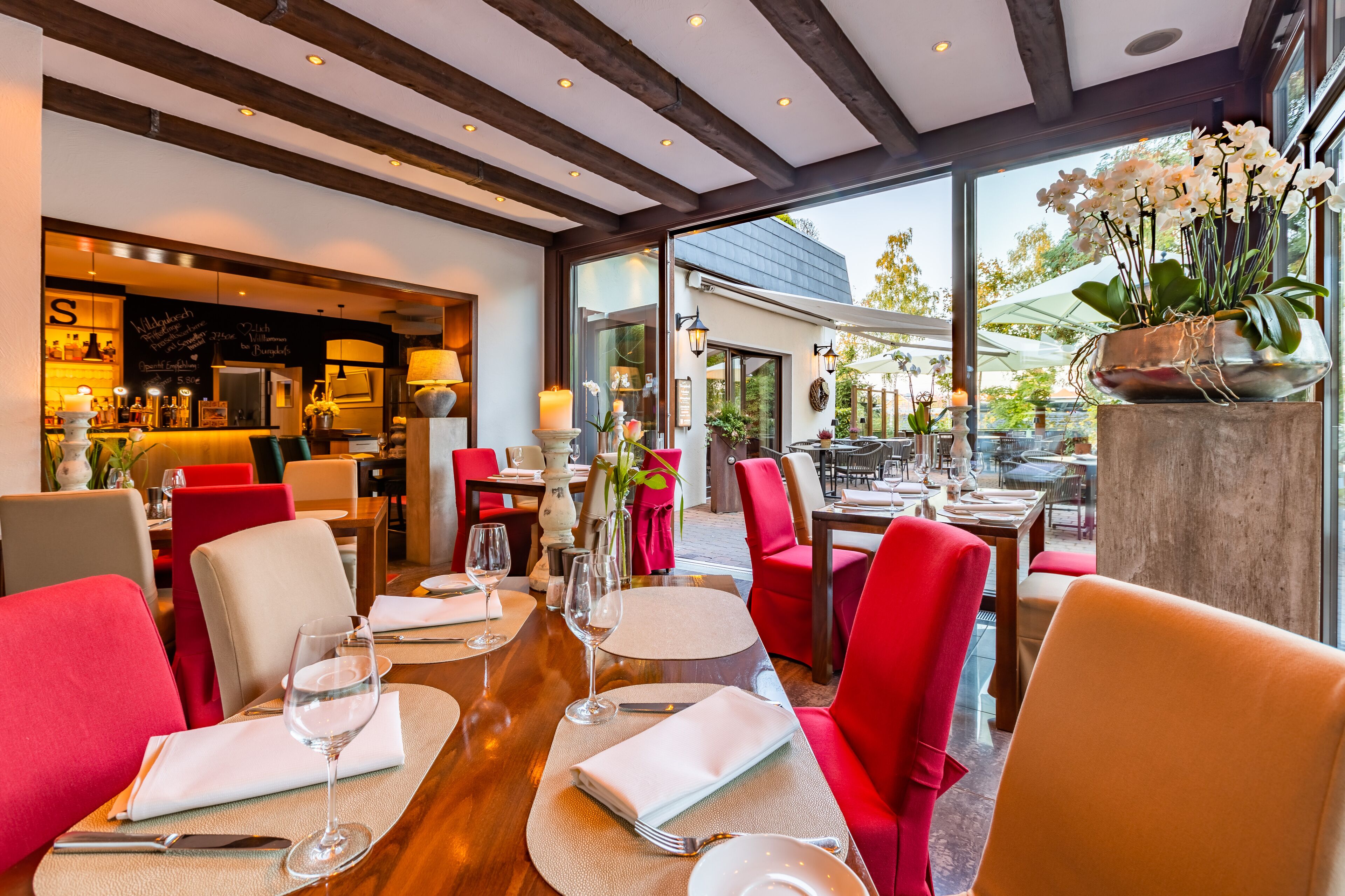 A restaurant with red chairs, round tables, and a bar area visible through glass doors.