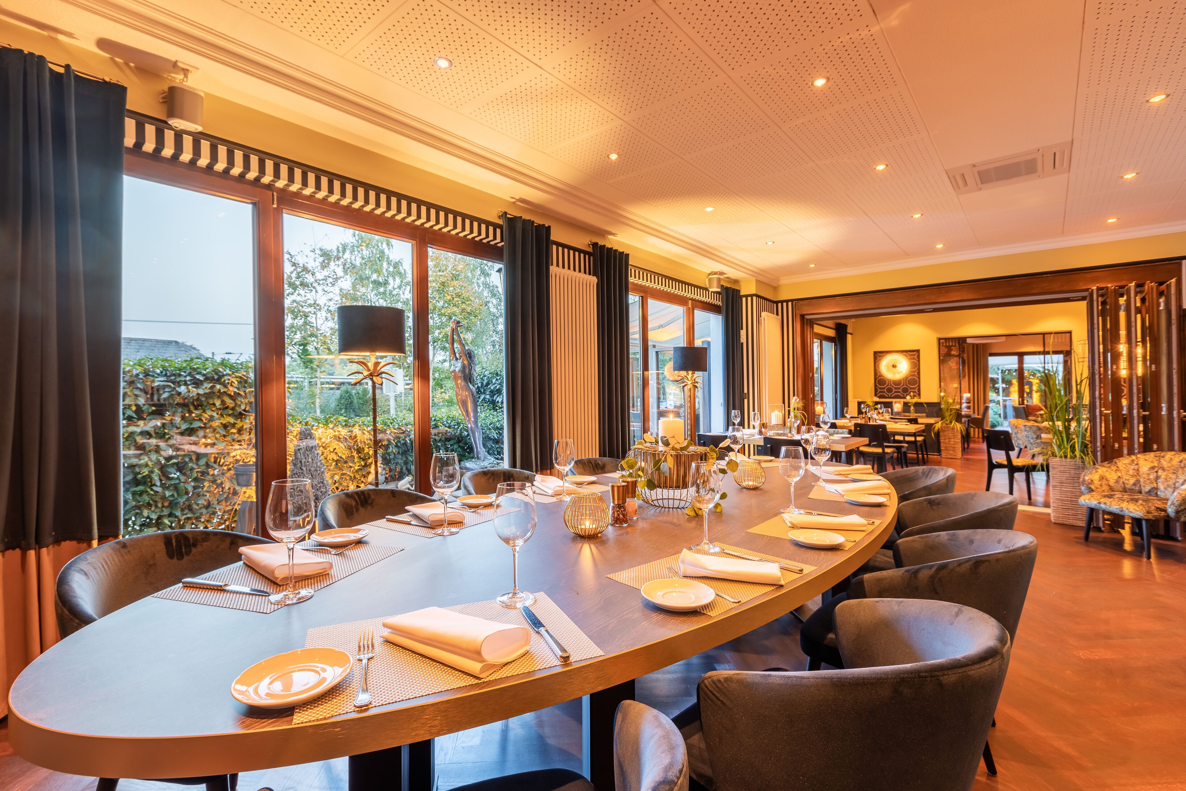 A dining area with a long table set for a meal, surrounded by chairs, with a view of greenery outside.