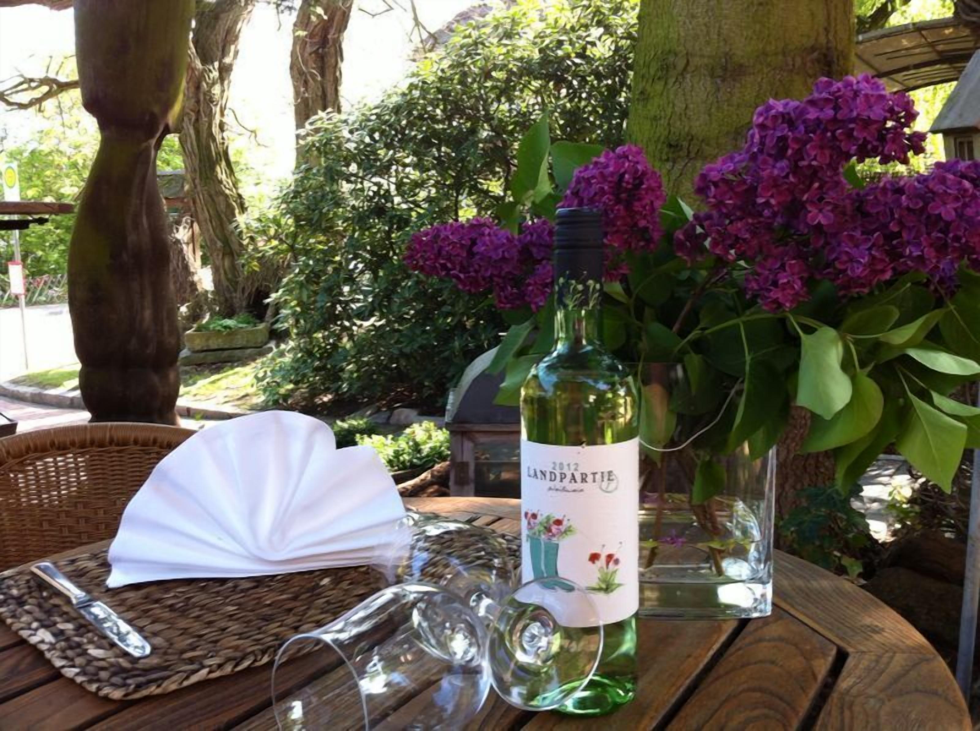 Una botella de vino, una copa, una servilleta y un jarrón con flores moradas sobre una mesa de madera al aire libre.