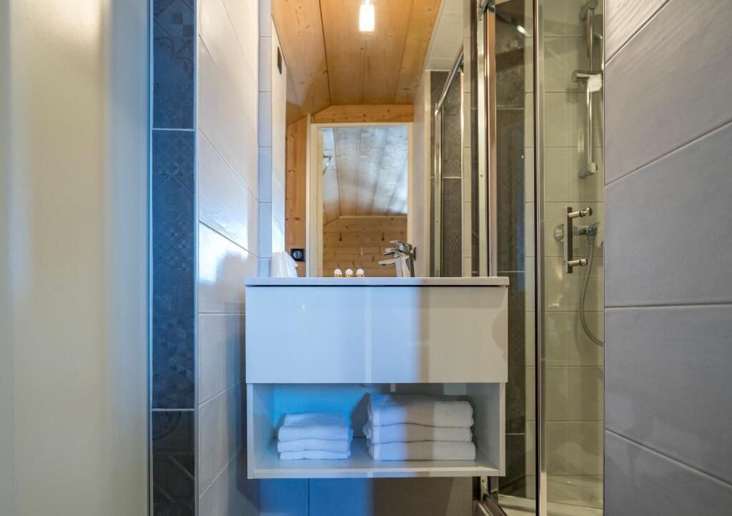A bathroom with a glass shower enclosure, a white sink with a mirror, and a shelf with folded towels.