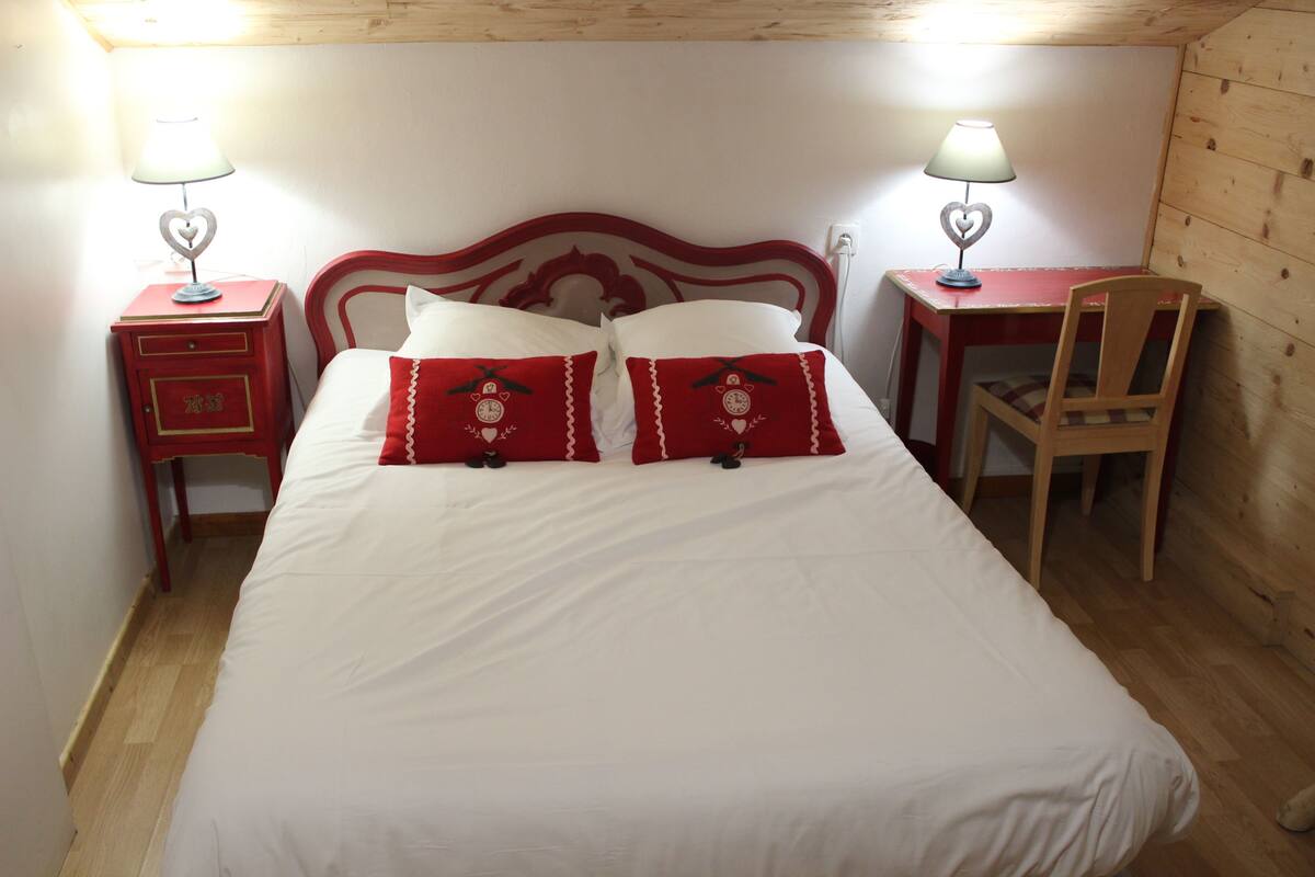 A bed with white bedding and red pillows, two bedside tables with lamps, a wooden chair, and a wooden desk.