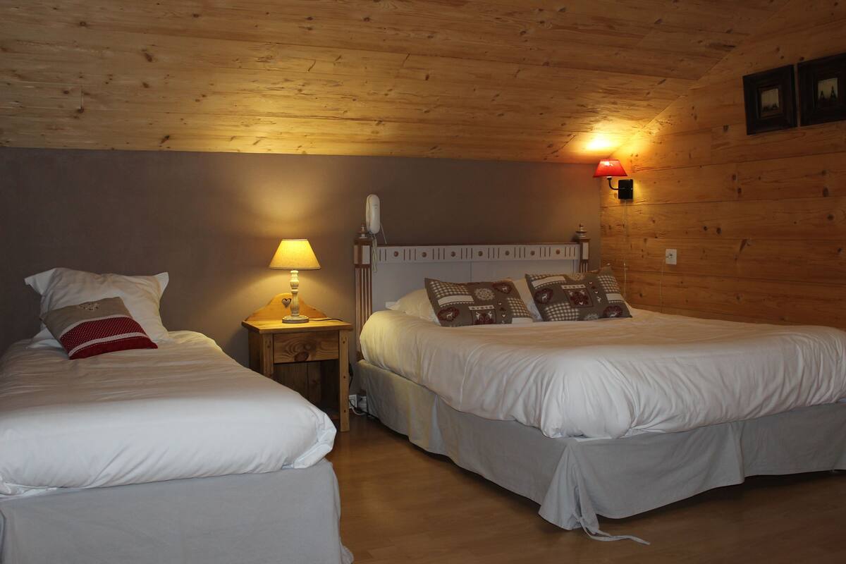 A room with two beds, wooden walls, a nightstand with a lamp, and a wall-mounted light.