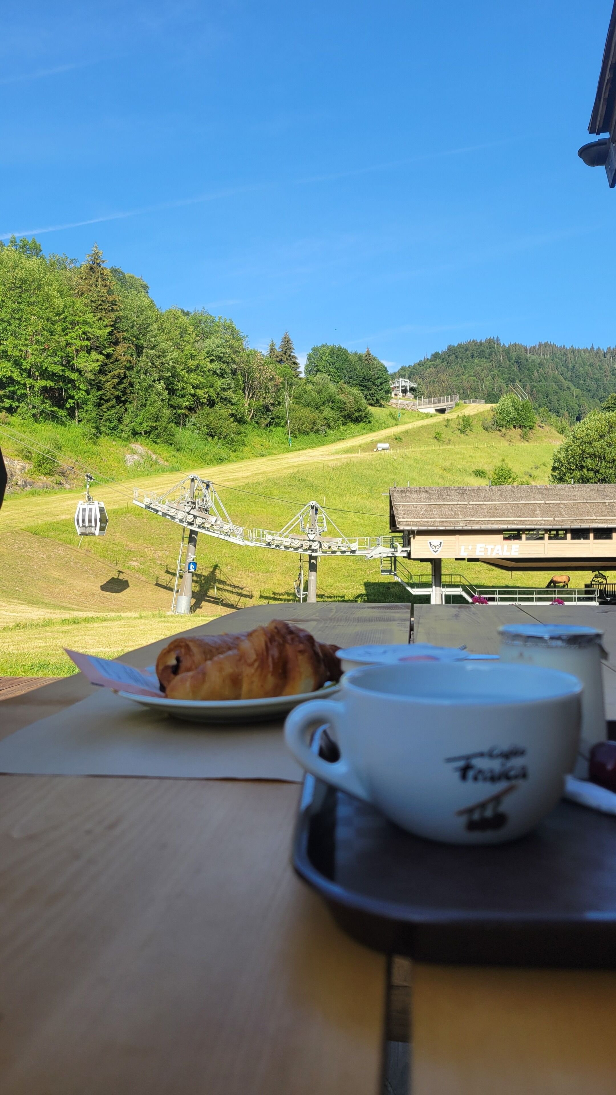 Une table avec un croissant et une tasse, donnant sur un téléphérique et un bâtiment portant l’inscription « L’Étoile », le tout dans un paysage verdoyant.
