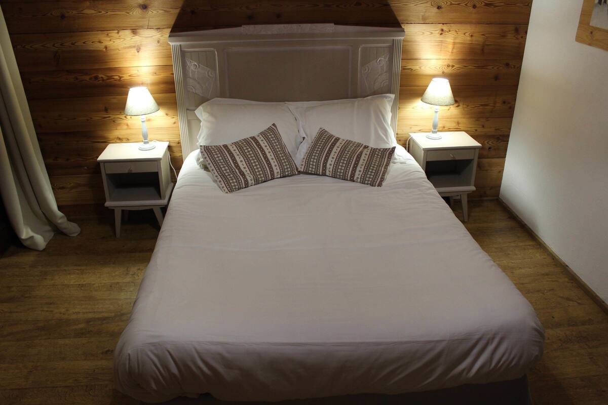 A neatly made bed with white bedding and two patterned pillows, flanked by two bedside tables with lamps, in a room with wooden paneling.