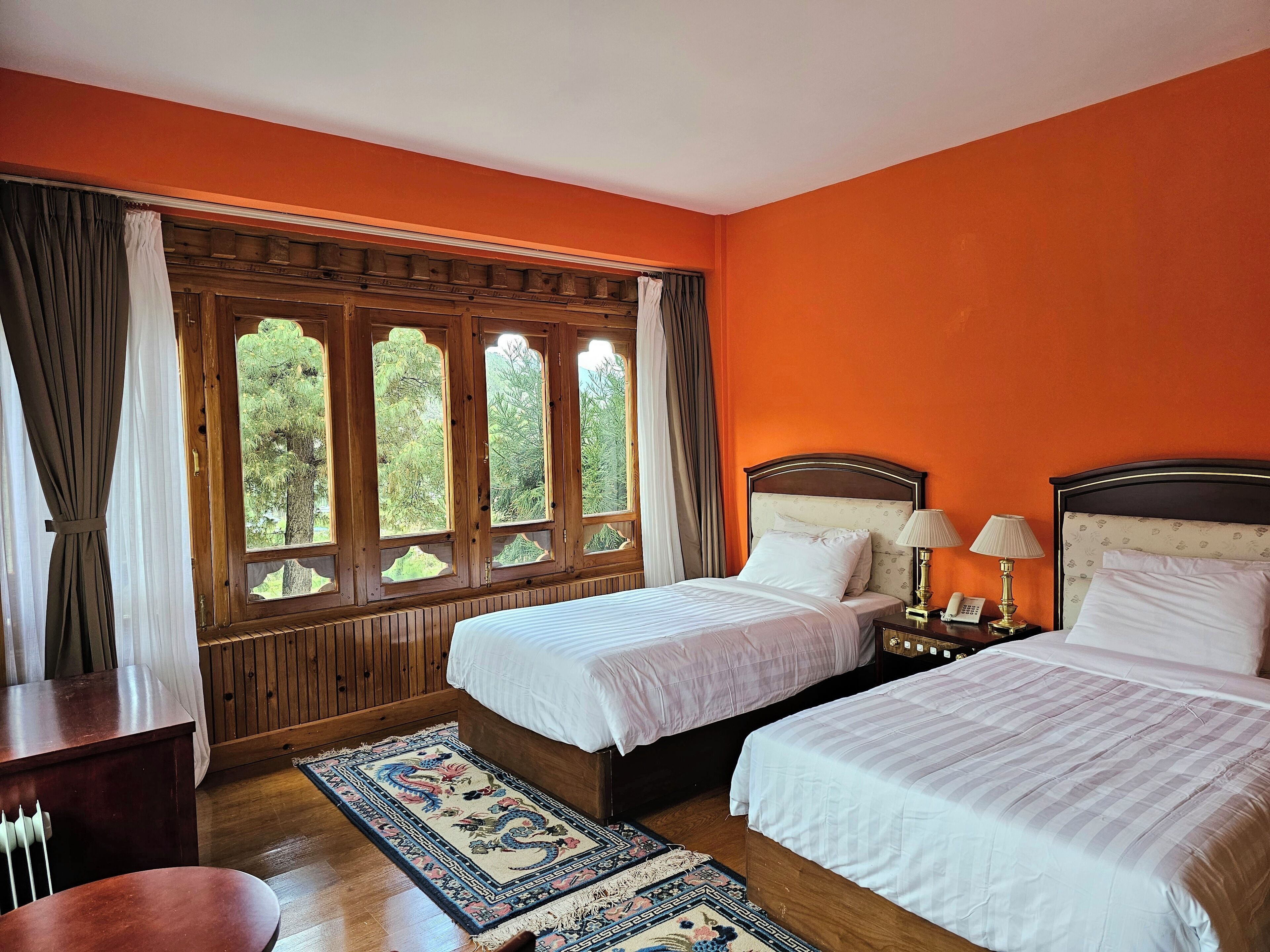 A hotel room with two beds, a wooden headboard, a nightstand with a lamp, and a view through a window with curtains.