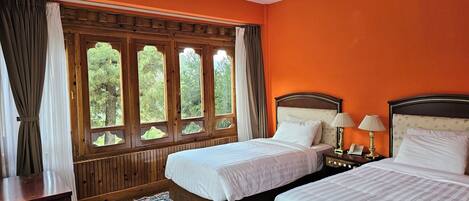 A hotel room with two beds, a wooden headboard, a nightstand with a lamp, and a view through a window with curtains.