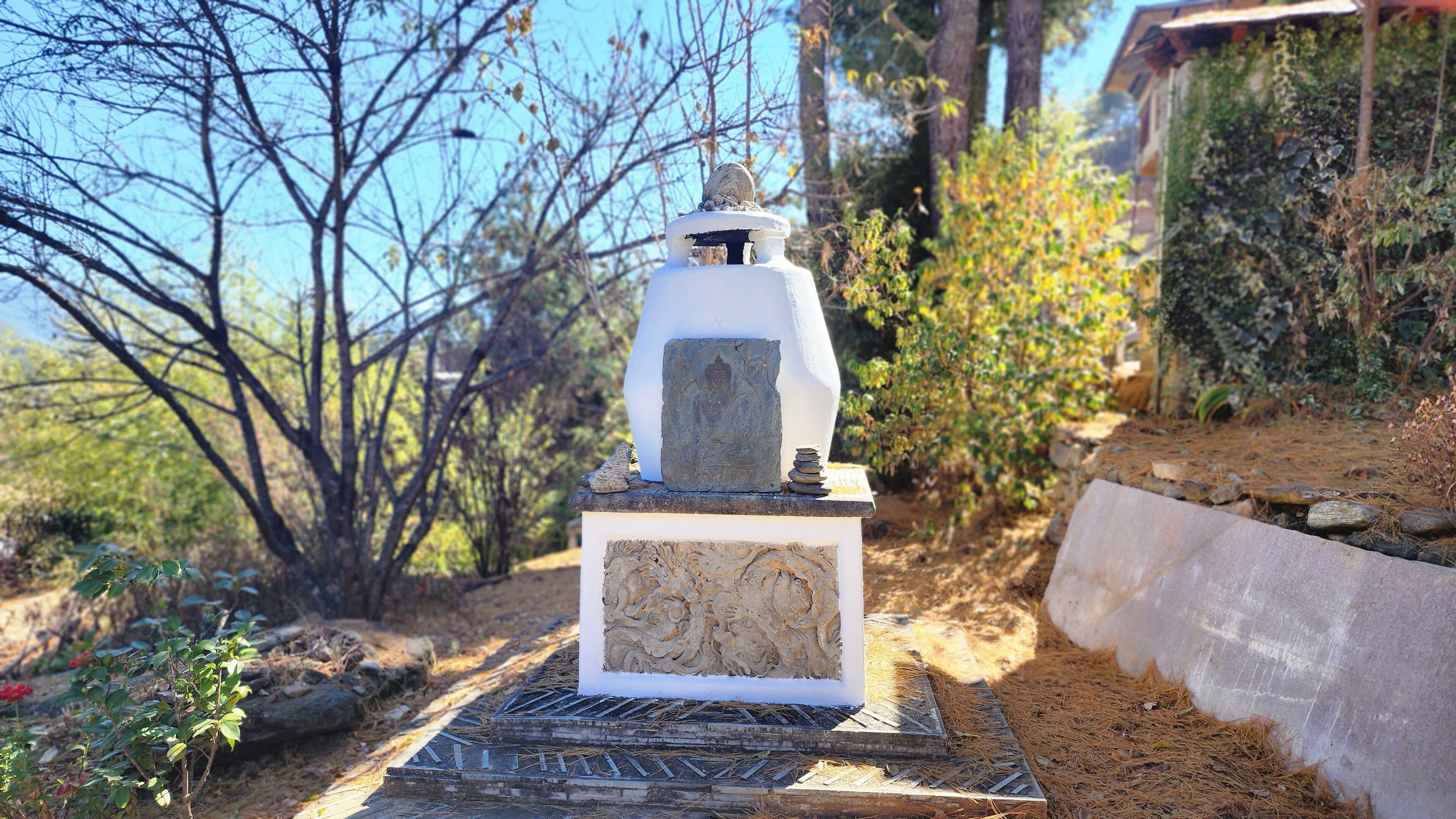 A white stone Buddha statue on a pedestal with intricate carvings, surrounded by natural vegetation.