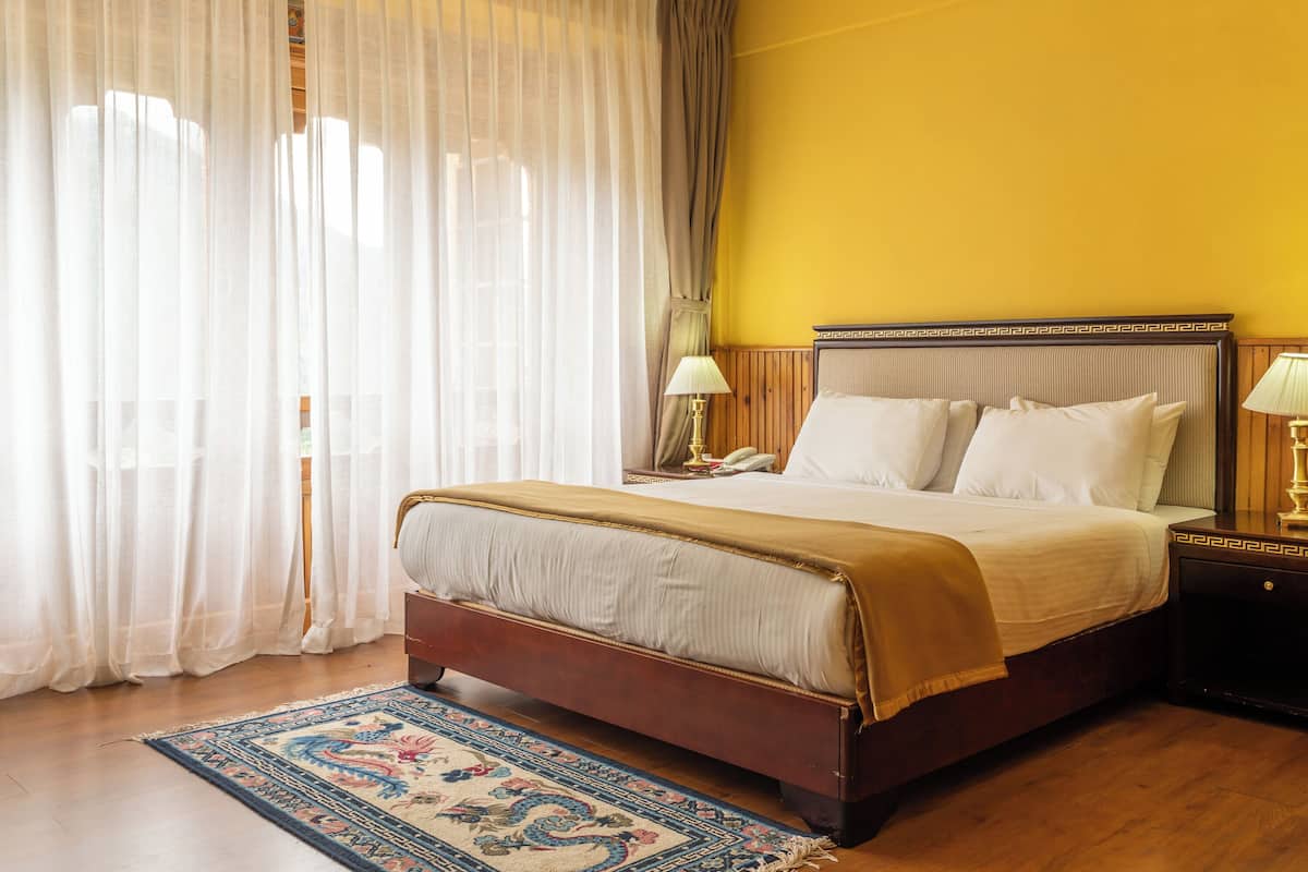 A hotel room with a large bed, a wooden headboard, two bedside tables with lamps, a patterned rug, and sheer curtains.