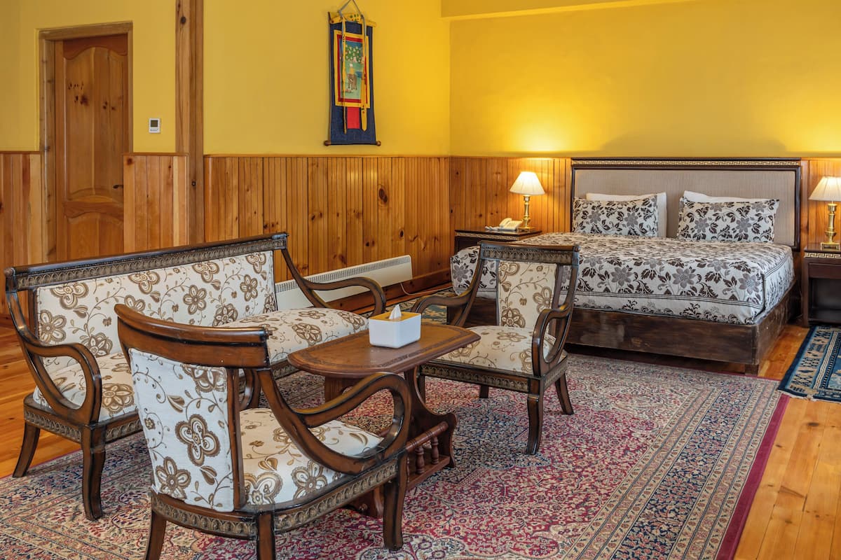 A room with a wooden paneled wall, a bed with floral bedding, two upholstered chairs, a small table, and a patterned rug.