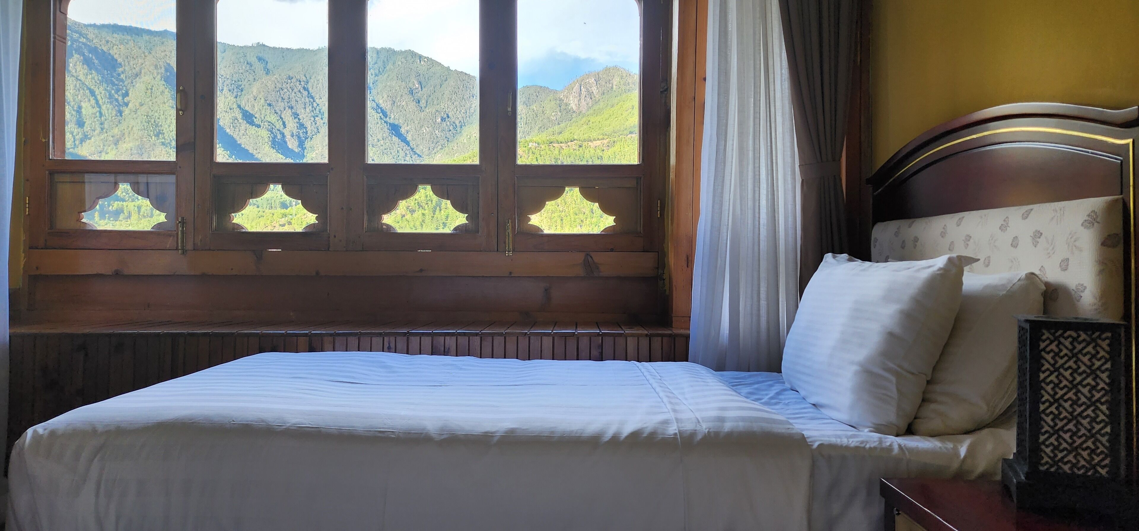 A bed with a wooden headboard and a view of green mountains through a window with decorative glass panes.