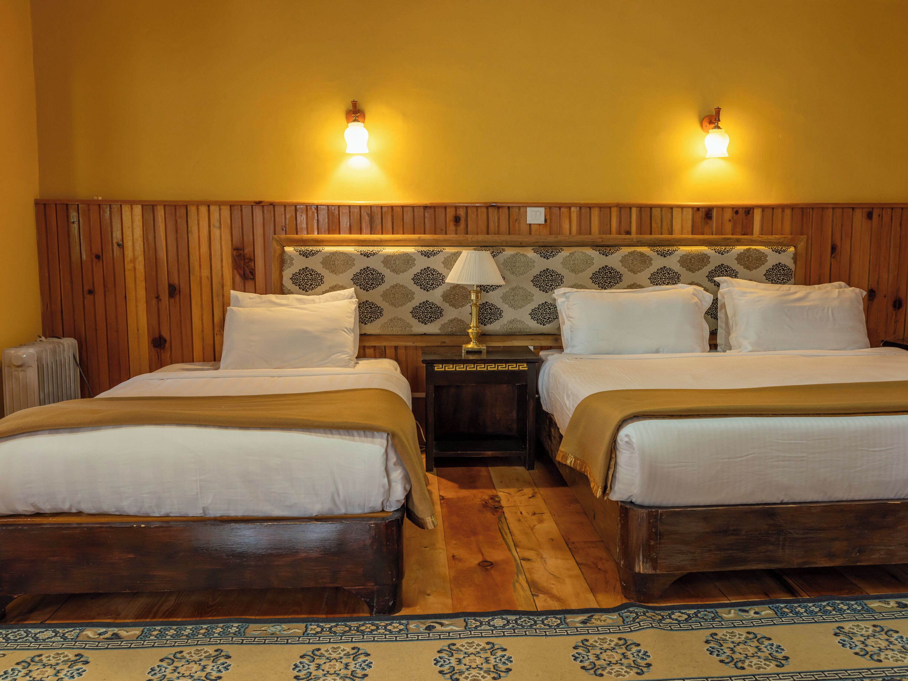 Habitación doble con dos camas, cabecero de madera, papel tapiz estampado, lámparas de noche y una alfombra decorativa.