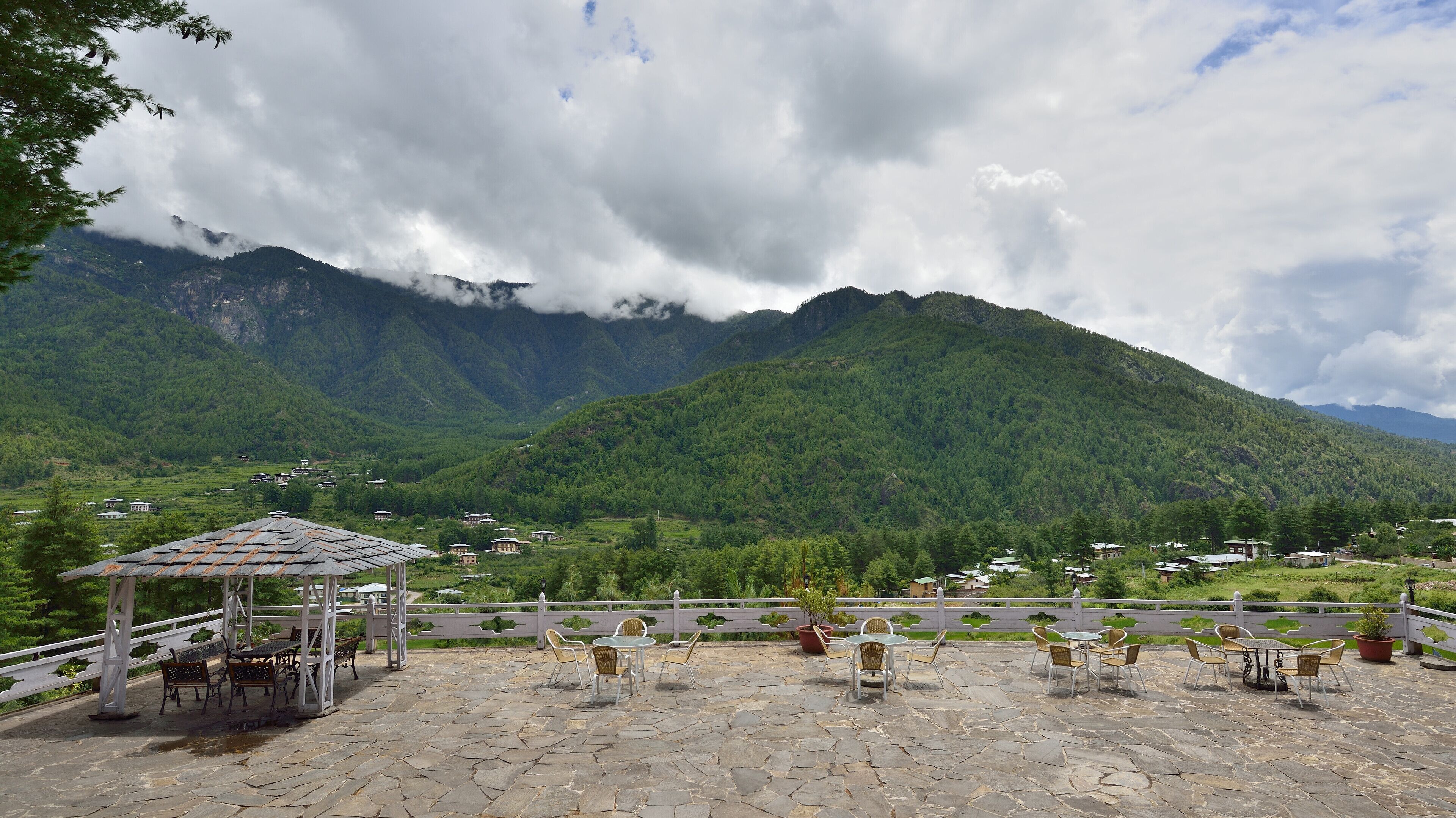 A stone patio with outdoor seating and a gazebo, overlooking a valley with mountains and scattered buildings.