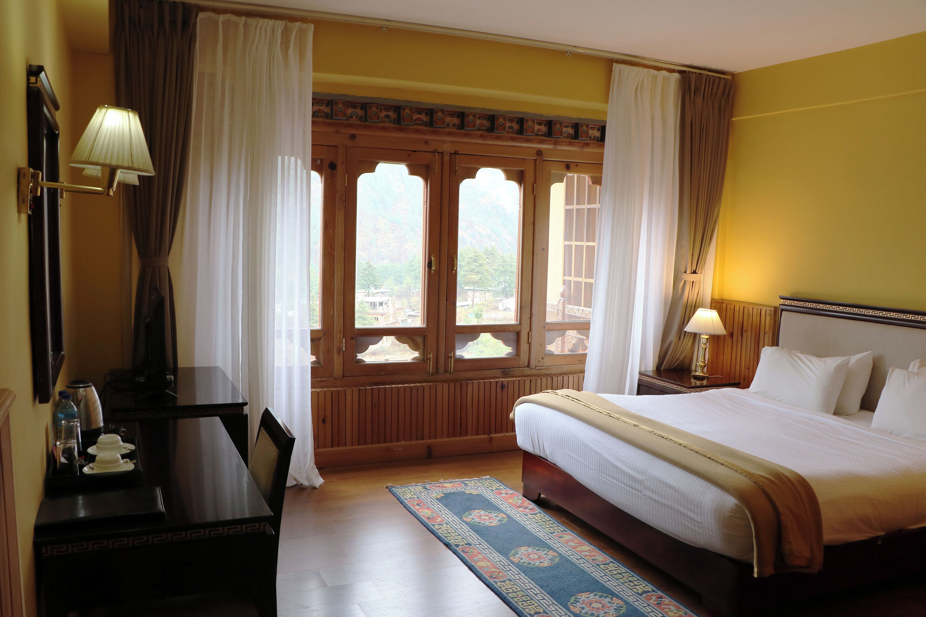 A hotel room with a bed, a desk with a lamp, a window with curtains, and a view of the outside.