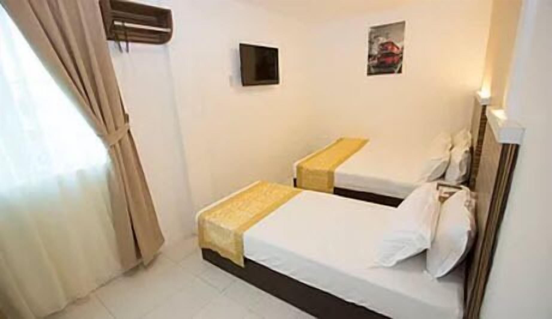 A small, clean room with two single beds, a small bedside table, a wall-mounted TV, and a window with curtains.